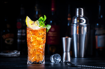 Boston tea cocktail drink with rum, gin, vodka, tequila, liquor, lemon juice, cola and ice in glass, dark background, bar tools and bottles