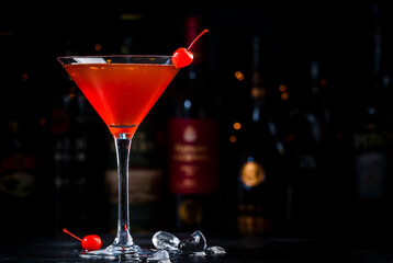 Manhattan cocktail drink with bourbon, red vemuth, bitter, ice and cherry in glass, dark bar counter background, bar tools and bottles