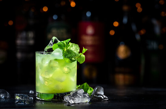 Green Negroni cocktail drink with dry gin, aperitif, gentian liqueur, fresh mint and ice. Black bar counter background, bar tools, bottles