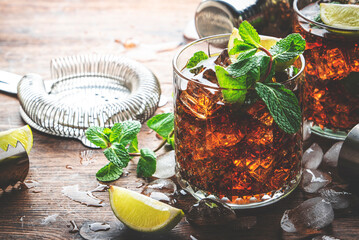 Rum cola cocktail drink with strong alcohol, mint, ice and lime in glass. Wooden background, hard...