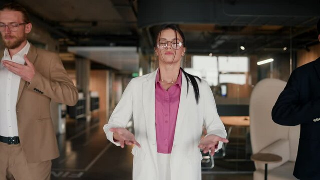Close-up shot of a brunette girl in round glasses in a white suit with a pink shirt doing breathing techniques with her colleagues during a break at work in a modern office