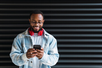 Happy smiling cool gen z young African American ethnic stylish hipster guy model standing at city urban wall using cell phone mobile device, looking at smartphone, holding cellphone. - obrazy, fototapety, plakaty