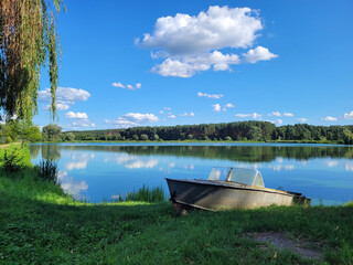 An old metal boat on the shore of a lake in the forest. clouds reflection in calm lake.  white clouds and a forest reflected