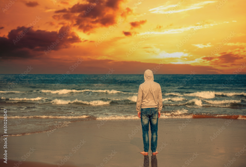 Wall mural Young man stands on the beach in the evening. The man gazing magical sunset - Wall murals