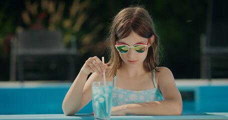 Cute little girl in sunglasses by the side of the pool. Nearby is a cool cocktail.