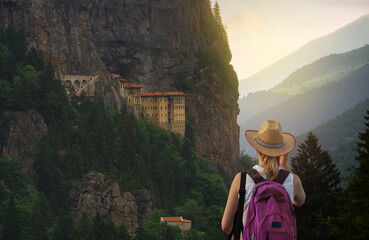 Tourist woman watches Sümela Monastery at sunset time. Sumela Monastery is one of the best tourist travel destinations in the Black Sea region of Turkey. Macka town, Trabzon , Turkey  - obrazy, fototapety, plakaty
