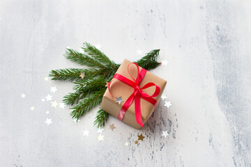 A small gift wrapped in kraft paper with a red ribbon on a light blue background with natural Christmas tree branches, surrounded by a shiny confetti star. Christmas background, holiday background