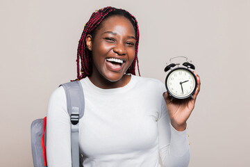 Delighted smiley African American woman with braids smiling holding alarm clock in hands showing at camera holding backpack on shoulders holsing clock in hands happy student.