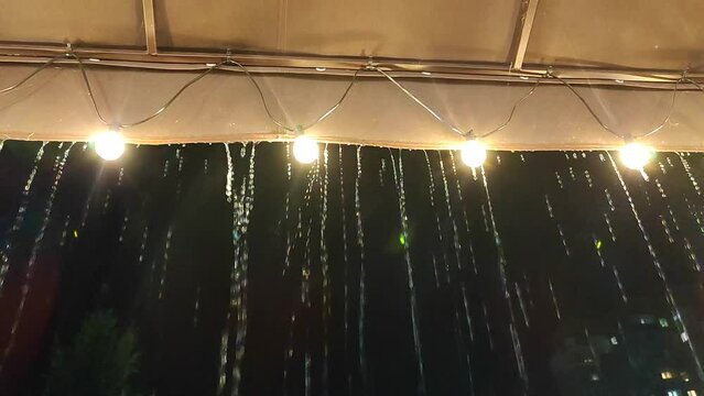 Streams of rain dripping from a roof with glowing lanterns at night. Streams jets of rain flowing from the edge of cover with garland of light bulbs on dark evening. Splashes and drops of heavy rain