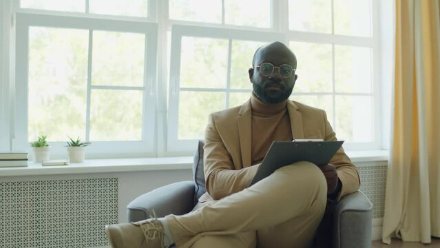 Medium long shot of African American psychologist sitting in armchair, writing notes on clipboard and posing for camera during workday in office