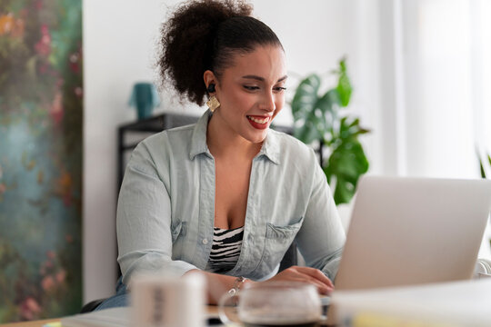 Side view of curved Middle Eastern female dressmaker in earbuds video calling on wireless laptop and working remotely at desk against blurred background
