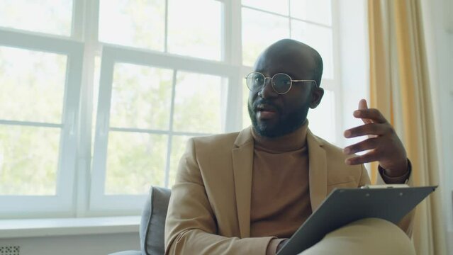 Medium shot of African American psychologist sitting with clipboard and giving advice to patient during therapy session