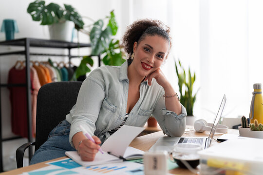 Portrait of optimistic Middle Eastern female seamstress with computer looking at camera and smiling while writing in organizer and working in showroom