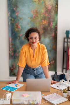Positive young female in casual clothes and makeup standing and leaning on table with laptop and chart while looking at camera and working on remote project