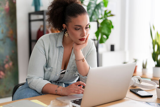 Middle Eastern female couturier in earbuds looking at screen of wireless computer and working remotely in workshop with rack of outfits