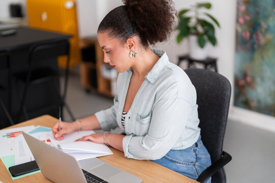 Side view of concentrated female curvy freelancer in smart casual sitting at table with laptop and writing notes in notebook while working on project