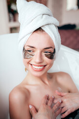 Portrait of young beautiful sexy woman having fun while lying in bathtub full of foam at home. Charming smiling model relaxing in luxury bath interior. Female with under eye patches. Towel on head