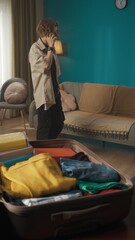 Obraz premium Vertical shot of a teenager, teen, young man having a call on the background while packing a suitcase laying on the foreground, discussing something.