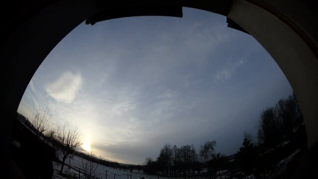 Window view with sky with sunrise to sunset, moving clouds, sun and halo, horizon with snow, trees and car traffic on two-lane road - timelapse, panorama 204.  Shortest day of year.