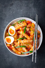 Tteokbokki with eggs, fresh green leek and roasted sesame seeds in blue bowl on the dark rustic table. Tteok-bokki is a Korean cuisine dish with rice gnocchi sticks and spacy sauce. Asian street food