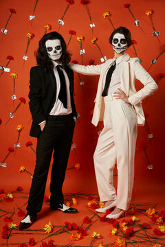 full length of stylish couple in skull makeup and suits standing on red backdrop with carnations