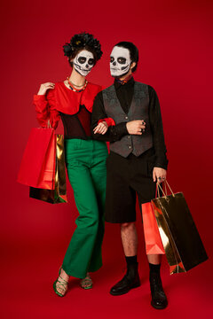 full length of couple in dia de los muertos makeup standing with shopping bags on red, festive sale