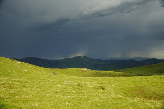 Paisaje de pradera verde bajo un cielo oscuro de tormenta con lluvia cayendo sobre las monta&ntilde;as.