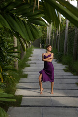 Outdoor portrait of stylish fashionable woman in purple silk dress in tropical resort. Natural light. Stylish, elegant summertime outfit, spring-summer fashion trend, tropical vacation style.