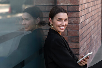 business woman in black office suit against brick wall talking at smartphone,smiling girl taking selfie or video call. mobile phone at ear, female support from window wall.outside street photo