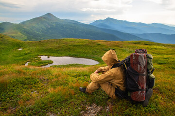 Tourist with a large backpack in a windproof suit in the mountains sits and looks at the mountain and mountain lake