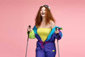 Beautiful happy ginger woman with stylish curly hair wearing stylish vintage overalls, holding ski equipment isolated on pink background. Travel, winter hiking concept 