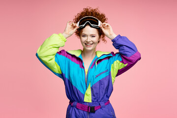 Smiling attractive red haired woman wearing stylish retro overalls, ski winter goggles looking at camera isolated on pink background. Travel, winter vacation, resort concept concept