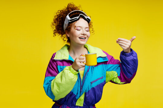 Attractive smiling woman wearing stylish overalls, ski goggles drinking coffee isolated on yellow background. Vacation, travel, winter resort concept