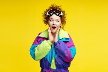 Portrait beautiful excited curly haired woman with open mouth wearing protective ski goggles,  winter overalls, looking away isolated on yellow background