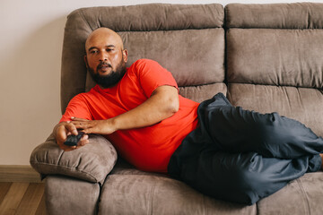 Fat and bald man watching TV while lying on sofa at home.