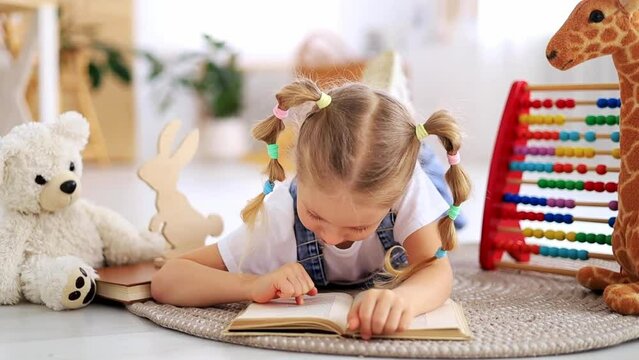 A Child Girl Reads A Book Lying On The Floor Among Toys In A Bright Bright Nursery Or Kindergarten, The Concept Of Children's Learning Centers Or Kindergartens And Learning