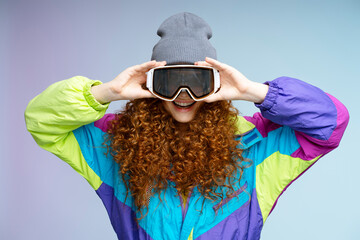 Portrait of beautiful smiling curly haired woman wearing ski goggles, winter hat isolated on blue looking at camera background. Vacation, winter,  travel concept