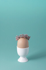 Light brown egg in white egg cup in wreath of pink flowers on pastel green background. Easter...