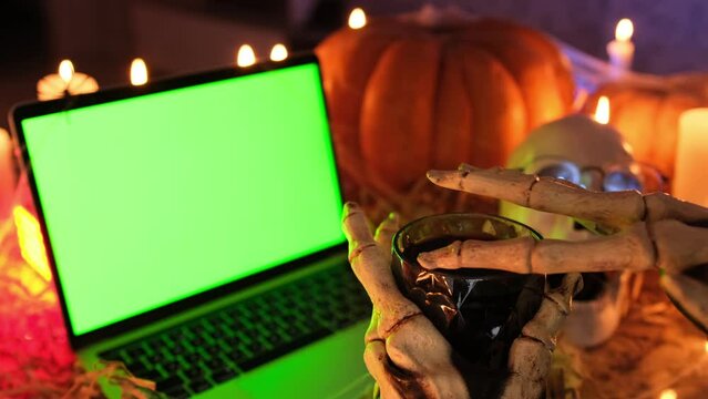 Creepy monster fingers stroke glass of blood near laptop with green screen among pumpkins and flickering candles in old dark crypt. Mysterious holiday background in yellow-red lamp light, side view.