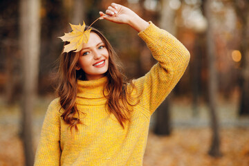 Young adult woman in autumn park holds in her hand yellow foliage maple leaf at face.