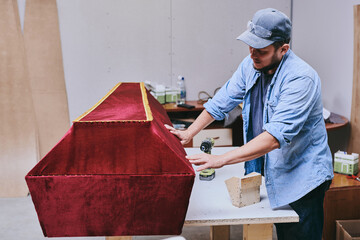 Carpenter works makes red velvet coffin in carpentry workshop. Man doing furniture professionally.