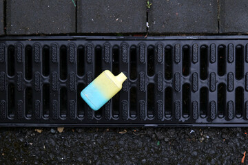A brightly colored e-cigarette vape has been discarded on a black plastic drain cover.