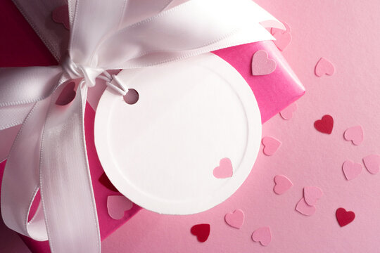 Empty white round gift tag with a heart on a pink gift box with a white bow. Space for text. Pink background.