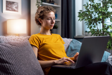 Young curly woman wear glasses using laptop to communicate with customer, or write email to office from home seating on couch in light room