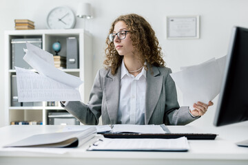 Cute beautiful woman reading notes, layouts, documents while sitting at desk in office