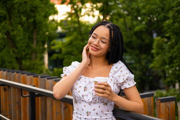 Naklejka premium African young attractive woman in glasses stands on a bridge with a paper cup of coffee on the street during the day in the park in summer