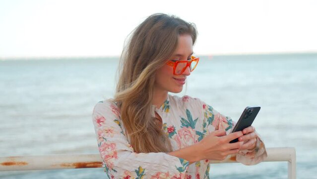 Caucasian woman tourist in beautiful dress and orange glasses standing leaning on railing on sea background and texting on cell phone. Using mobile technologies during summer vacation