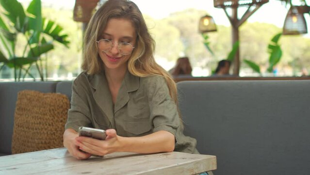 Young beautiful blonde woman using smartphone while enjoying summer on outdoor terrace cafe or coffee shop. Stylish female person wear trendy glasses showing teeth smile holding mobile phone in hand