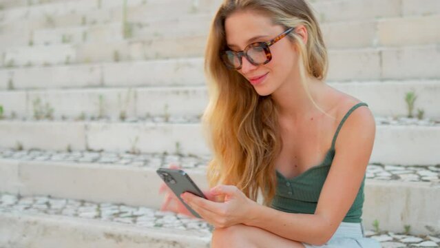 Pretty young woman texting on cell phone sitting on stairs outdoor. Happy girl chatting online during free time on city street. Woman looking into camera, correcting glasses and smiling