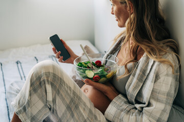 A pregnant woman sitting in pajamas eats salad and uses a mobile phone to communicate.